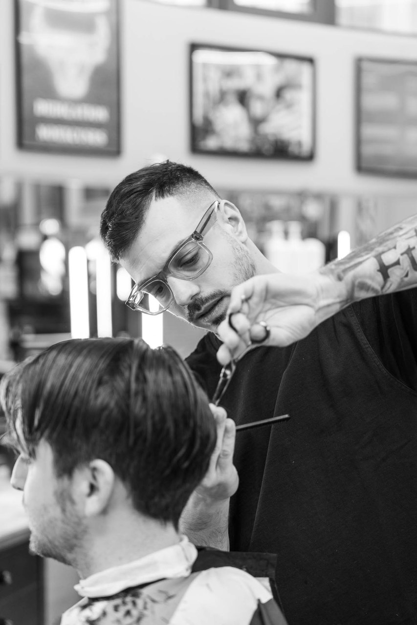 Un barbiere con gli occhiali taglia con le forbici, in primo piano i capelli del cliente