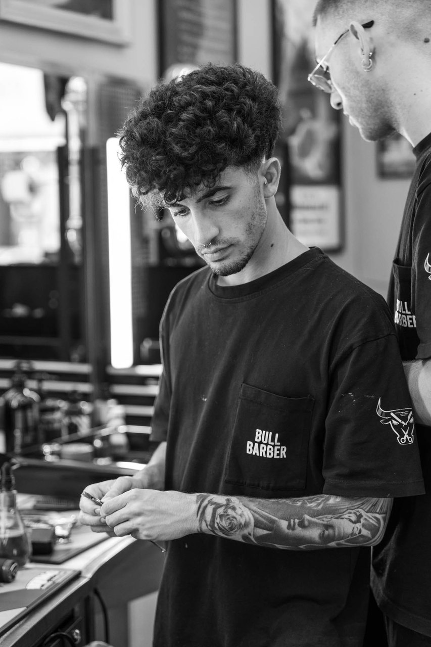 Un barbiere riccio con la maglia Bull Barber sistema il rasoio fra le mani, alla postazione