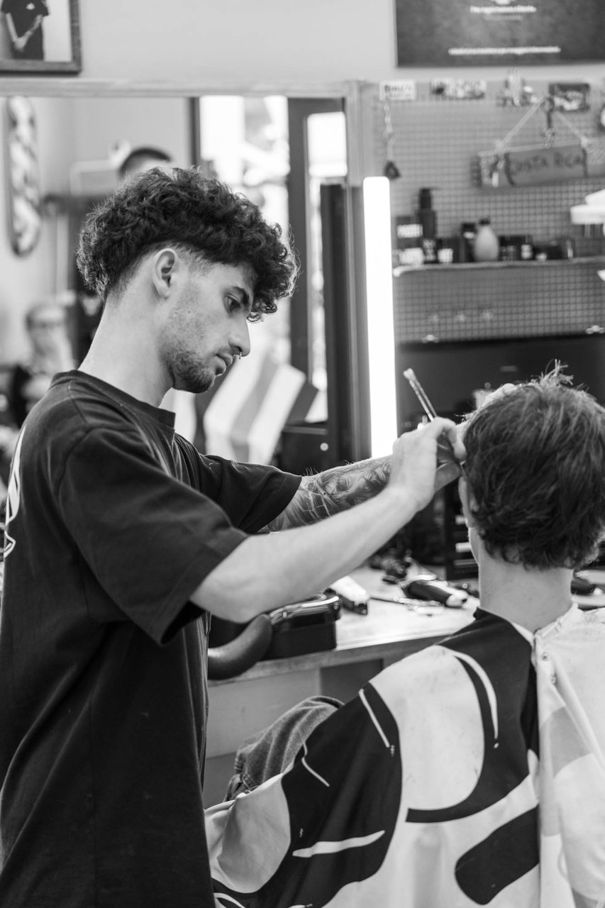 Un barbiere riccio taglia con le forbici i capelli di un cliente di spalle, davanti allo specchio