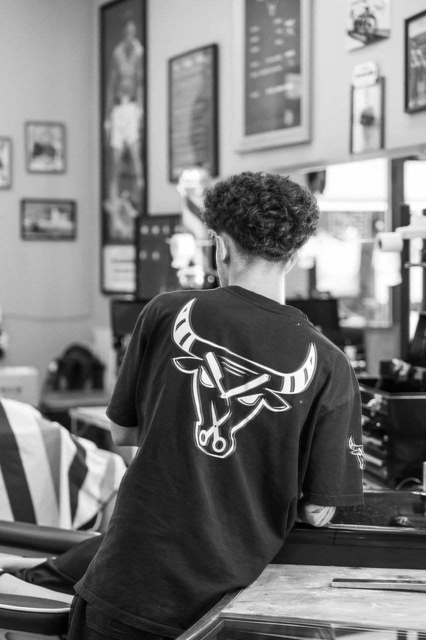 Un barbiere di spalle appoggiato alla postazione, il toro sulla maglia e i quadri alle pareti