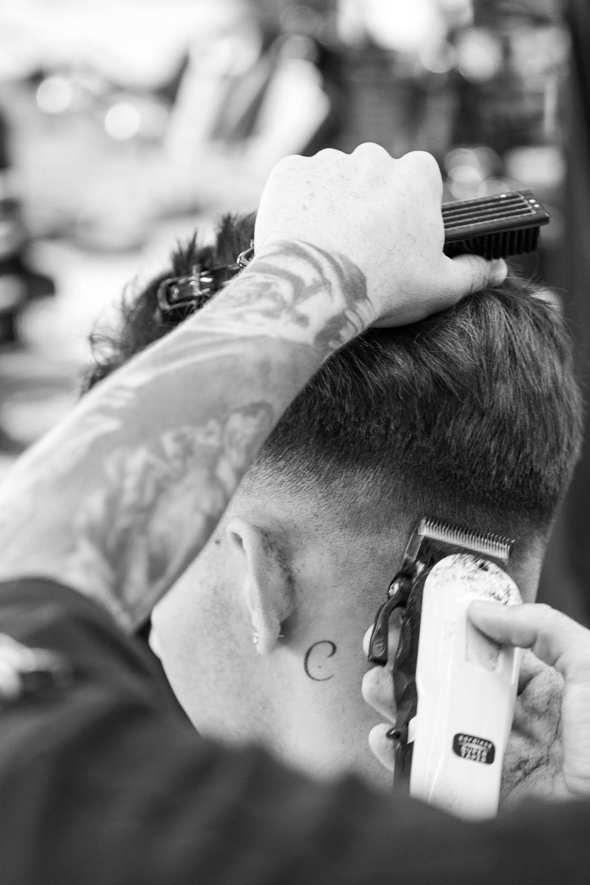 La macchinetta sfuma la nuca mentre la mano tatuata tiene su i capelli col pettine