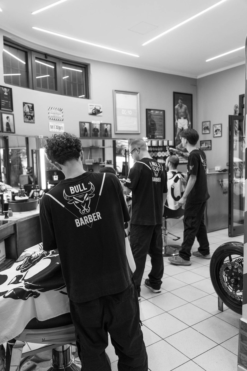 La sala di Bullbarber a Montale: tre barbieri di spalle al lavoro, col toro sulla maglia, le postazioni e le finestre alte
