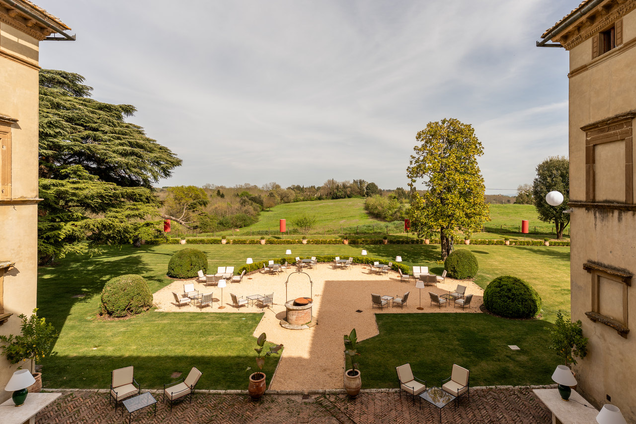 Il giardino visto dall'alto fra le due ali della villa: il pozzo, la terrazza in ghiaia coi tavolini, il prato, il cedro e la campagna