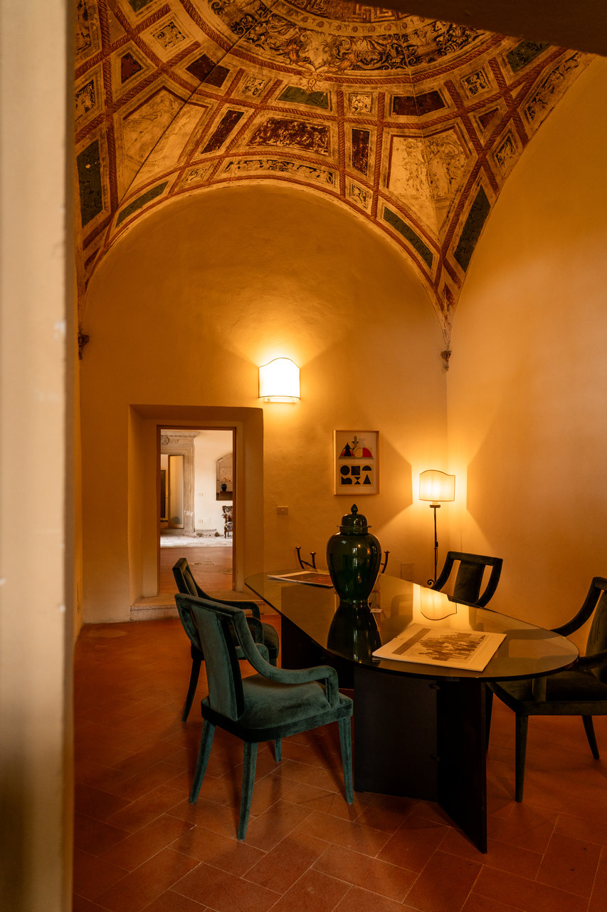 La sala con la volta a cassettoni dipinti, il tavolo di vetro e le sedie di velluto verde, la porta aperta sulle stanze accanto
