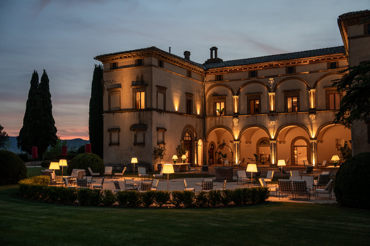 La villa di tre quarti all'ora blu: il loggiato del cortile acceso, la terrazza in ghiaia coi tavolini e le lampade, i cipressi e il cielo rosa