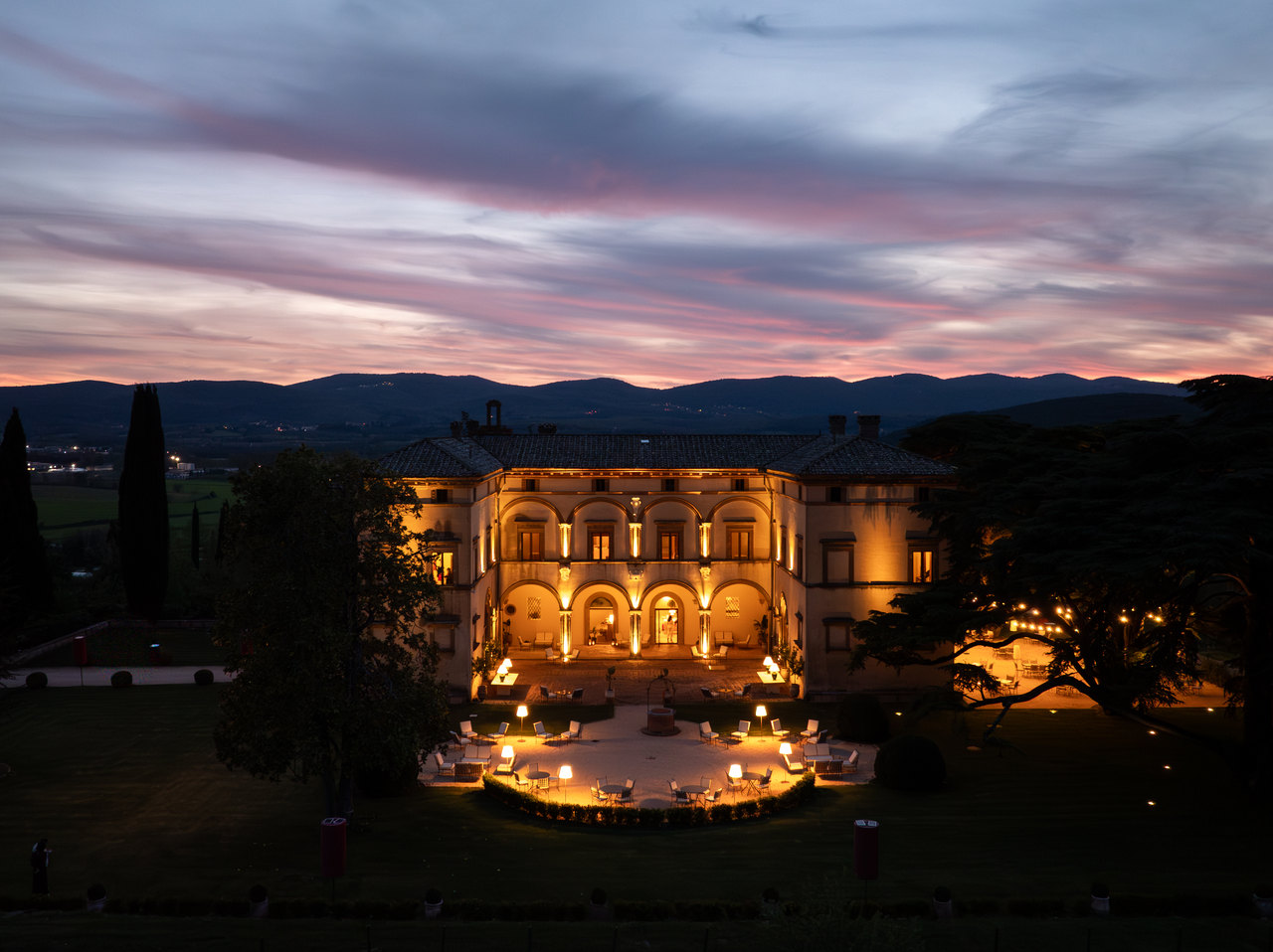 La villa all'ora blu vista dal drone: il cortile con gli archi su due piani illuminato, la terrazza a semicerchio con le lampade accese, il cedro e il cielo rosa sopra le colline