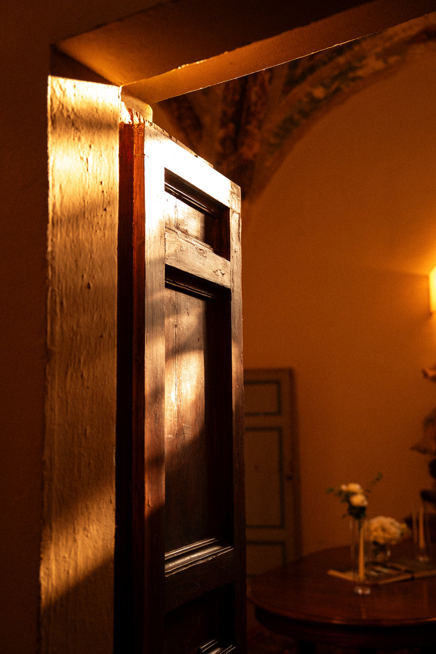 Una porta di legno socchiusa nella luce radente del pomeriggio, oltre la soglia un tavolino coi fiori