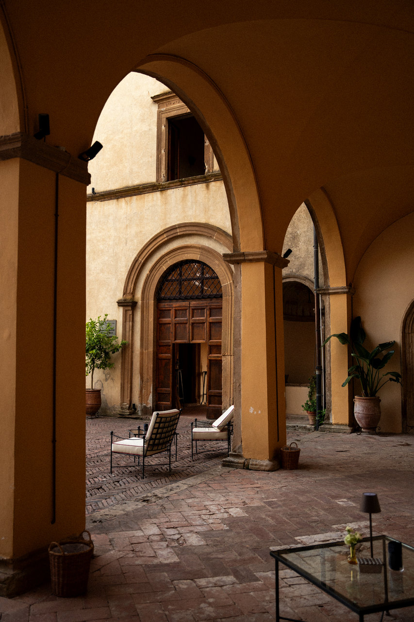 Sotto il loggiato del cortile: gli archi, il portale di pietra con la lunetta, le poltroncine e un tavolino di vetro, nella luce del tardo pomeriggio