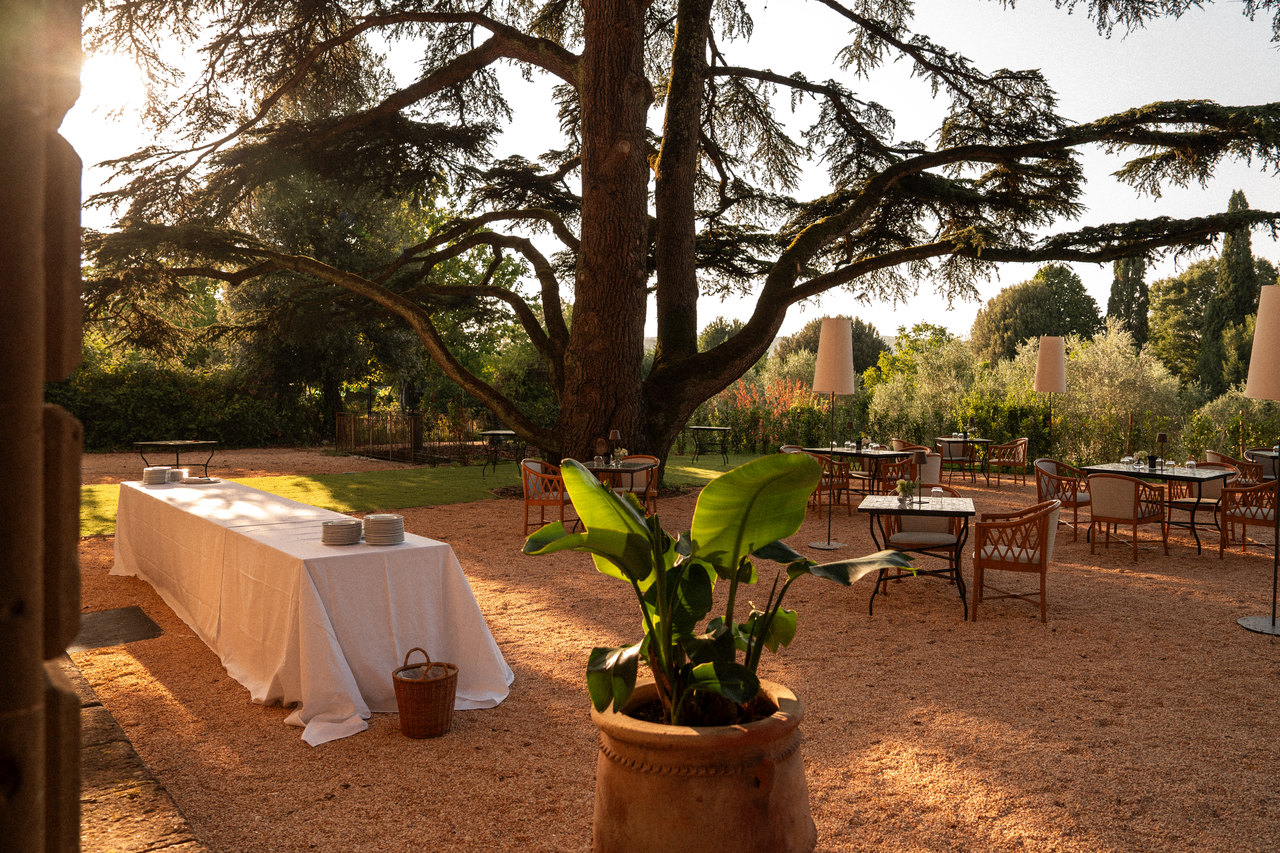 La terrazza sotto i cedri al tramonto: il tavolo lungo del buffet con la tovaglia bianca, i tavolini e le lampade