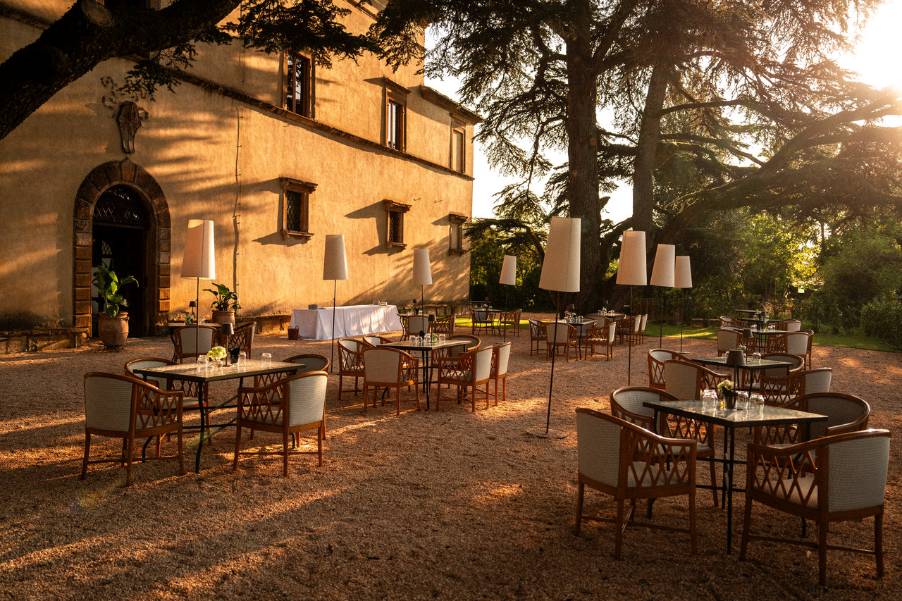La facciata laterale col portale nella luce del tramonto, i tavolini e le lampade alte sulla ghiaia sotto il cedro